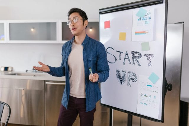A young man in a blue jacket gestures as he speaks next to a whiteboard displaying the word "STARTUP" and colorful sticky notes, in a modern office space.