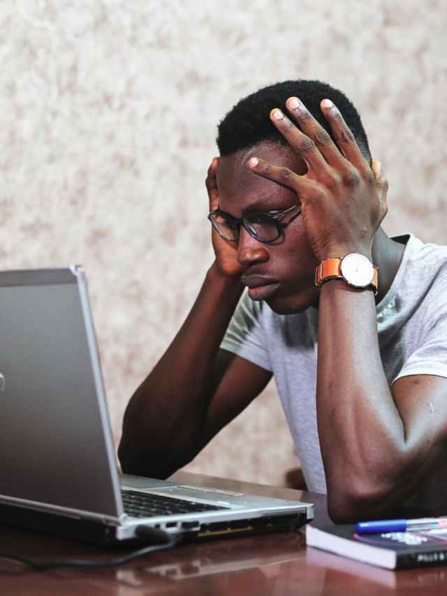 A young man with glasses sits at a desk, resting his head in his hands, looking stressed while staring at a laptop screen. A notebook and pen are visible beside him.
