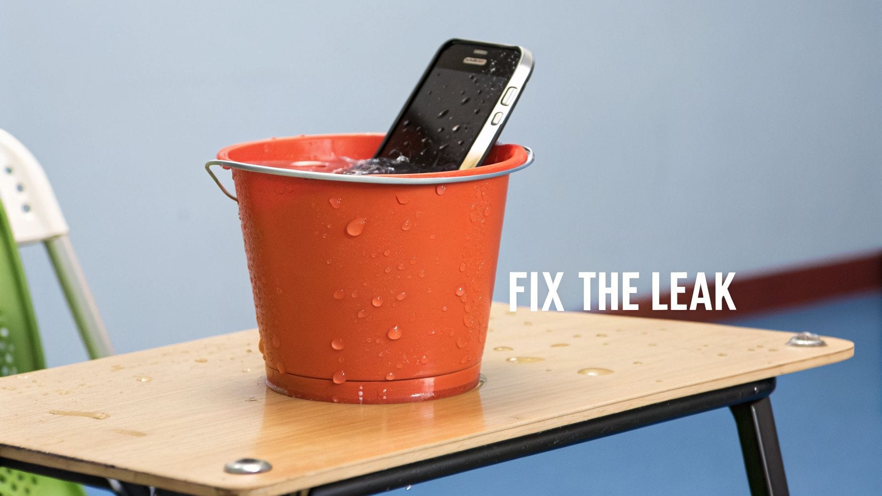 A black smartphone is partially submerged in an orange bucket filled with water, placed on a wooden table. Water droplets are visible on the bucket's surface, with the text "FIX THE LEAK" be
