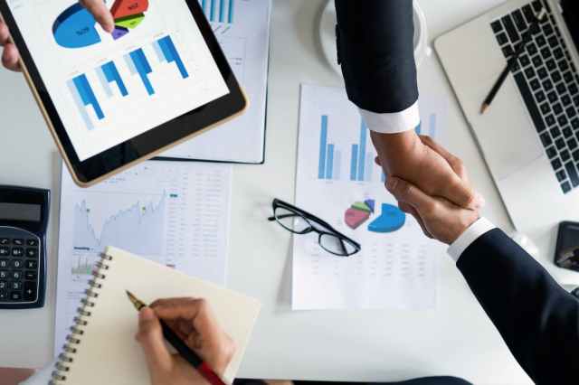 Two business people shaking hands over a desk filled with documents and graphs, a tablet displaying charts, a notebook, a calculator, and glasses present.