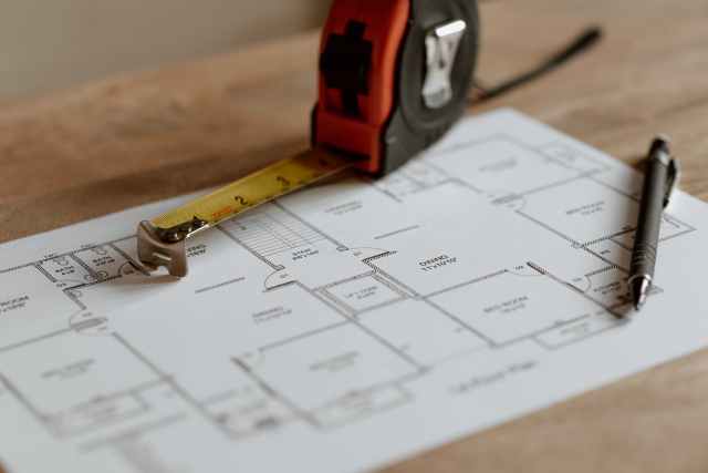 A tape measure and a pen rest on a floor plan diagram placed on a wooden table, illustrating a layout with various labeled rooms and spaces.