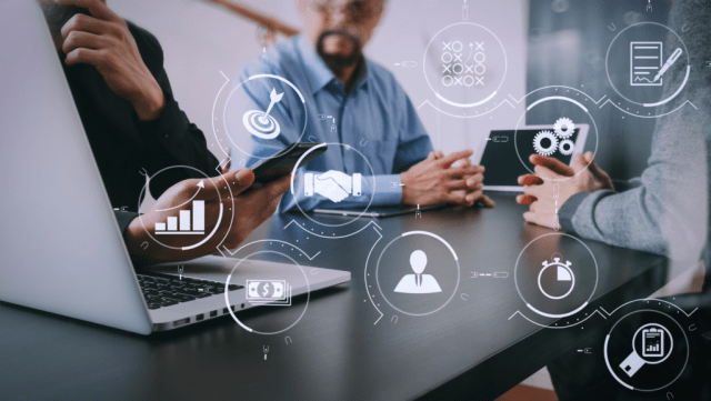 Three people are seated around a table with a laptop open, discussing ideas. Various icons representing analytics, teamwork, and deadlines are overlaid in a digital style.
