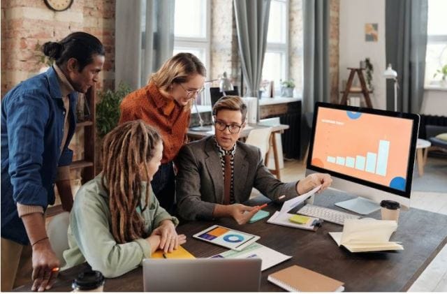 A diverse group of four people collaborates in a bright, modern office. They analyze documents and a computer screen displaying a bar graph and sectioned colors on an orange background.