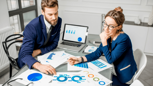 A man and woman in business attire are reviewing documents with charts and graphs on a table. A laptop displaying analytics is open in the background. Bright, modern office setting.
