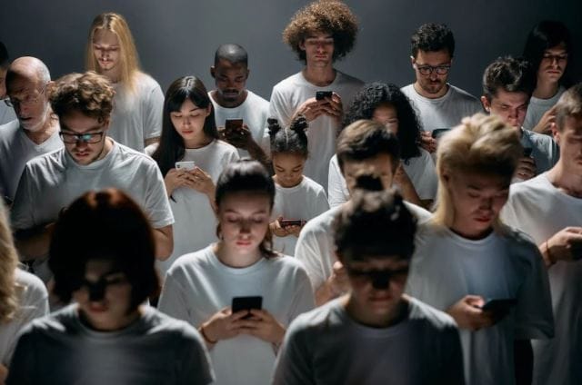 A diverse group of people in white shirts, focused on their phones. They stand in a dimly lit space, with a mix of genders, ages, and hairstyles, reflecting engagement with technology.