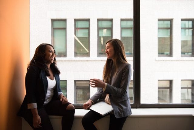 Two women are sitting on a ledge in a bright office space, smiling and engaging in a conversation, with large windows and a view of buildings in the background.