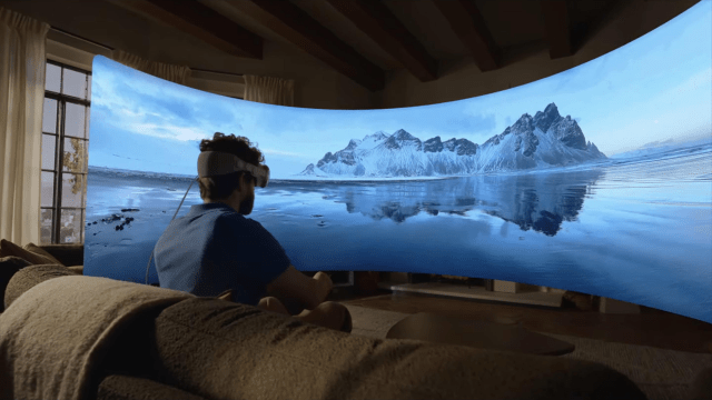A person wearing a headset sits on a couch, facing a large, curved screen displaying a serene landscape of mountains reflecting in water, with a cozy, warmly lit room in the background.