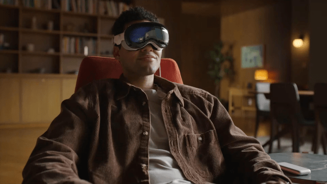 A man sits in a comfortable chair wearing Apple Vision Pro goggles, looking relaxed and focused. The room features wooden shelves, a plant, and soft lighting in the background.