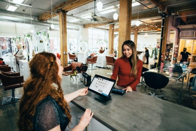 A stylist and a customer engage at a sleek salon counter, with a tablet displaying information between them. Chairs and mirrors fill the bright space in the background.