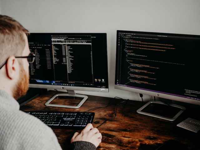 A person with a beard works at a wooden desk, focused on two monitors displaying lines of code and file directories, with a black keyboard in front.