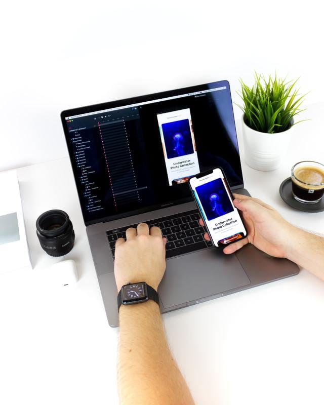 A person using a laptop with a coding interface, holding a smartphone displaying a mobile app design, surrounded by a plant, coffee, and a camera lens.