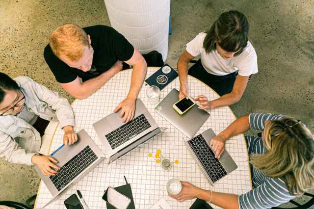 A group of four people working together at a circular table with laptops and a smartphone, engaged in discussion and collaborating on app performance.
