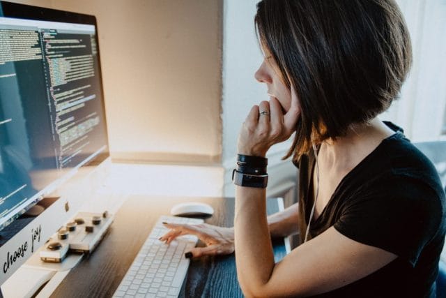 A woman with short hair sits at a desk, looking intently at a computer screen displaying lines of code. She has one hand on her chin and wears several bracelets.