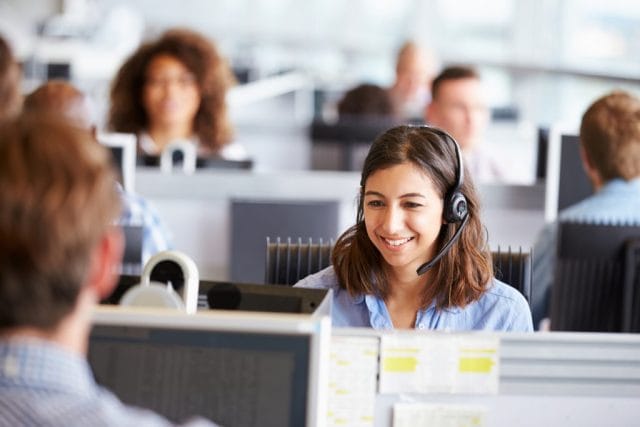 A smiling customer service representative wearing a headset sits at a desk, with colleagues visible in the background at their workstations in a busy office environment.
