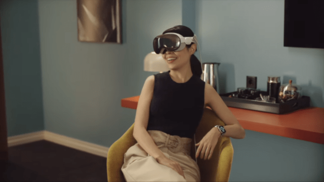 A smiling woman wears sleek virtual reality goggles while sitting in a stylish chair. She has short hair and is dressed in a black top and beige skirt, with a watch on her left wrist.