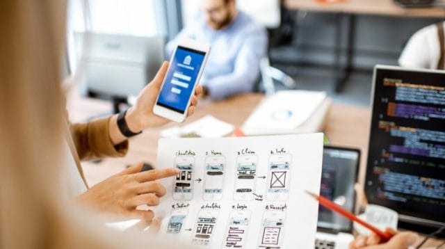 A person points to a printed sheet displaying mobile app design sketches while holding a smartphone showing a login screen. In the background, team members work at desks with laptops and cod