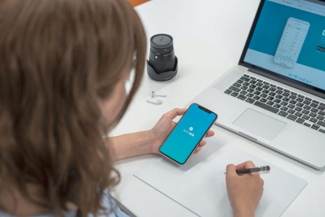 A person with brown hair is holding a smartphone displaying a blue screen while writing notes on a blank page. A laptop and a camera are visible on the table.