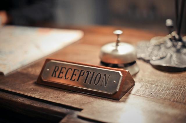 A close-up of a wooden reception desk featuring a metal nameplate that reads "RECEPTION," alongside a small bell and an ornate object. A blurred map is visible in the background.