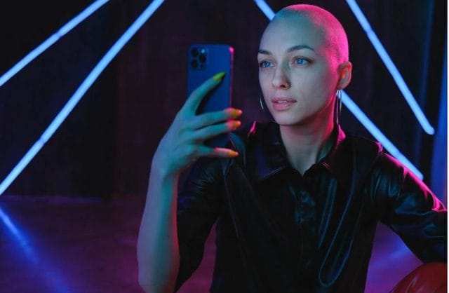 A person with short hair and bright nails holds a smartphone while taking a selfie, surrounded by colorful neon lighting in a dark setting.
