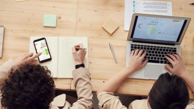 A person using a smartphone displays a graph, while another person works on a laptop with colorful data charts. A notebook, pen, and sticky notes are on a wooden table.