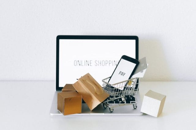 A laptop displaying "Online Shopping" with a small shopping cart filled with boxes and a smartphone showing the word "PAY" next to it, representing e-commerce.