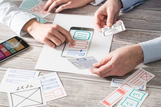 Two hands manipulate paper app interface mockups around a smartphone, which displays a design layout, all set on a wooden table. A smartphone with colorful app icons is in the corner.