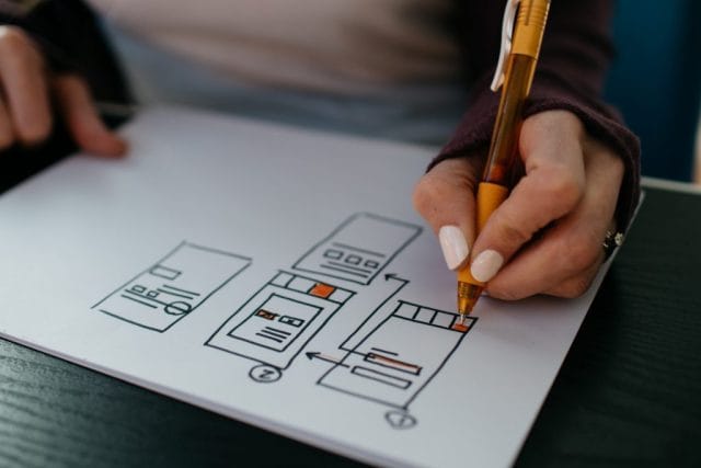 A person with a pen sketches app design concepts on a notepad, featuring simple diagrams of mobile screens and user interface elements, on a dark wooden table.