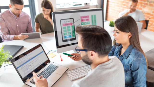 A diverse team collaborates in a modern workspace. Two people discuss app design while using a laptop, with a large screen displaying a mobile app interface in the background.