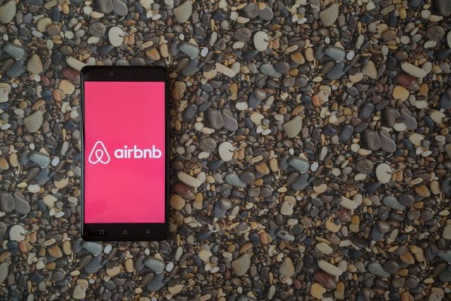 A smartphone displays the Airbnb logo on a vibrant pink background, placed atop a textured surface of small, colorful pebbles.