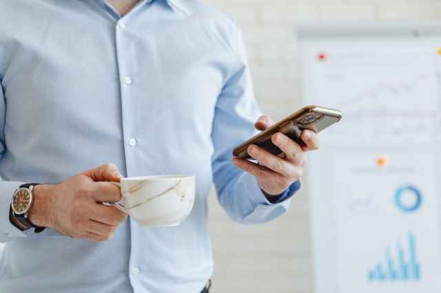 A person holding a smartphone in one hand and a white marble coffee cup in the other, wearing a light blue shirt. A blurred presentation board with graphs is visible in the background.