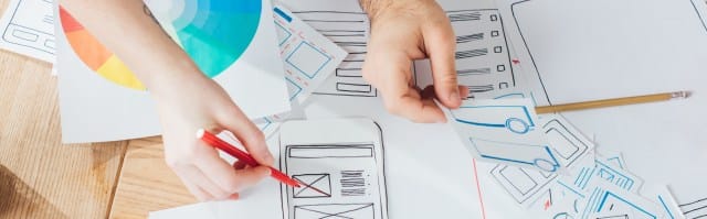 Hands sketching app layouts and wireframes on papers scattered on a wooden table, with a color wheel in the top left corner. A pencil and red pen are visible among the sketches.