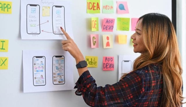 A woman points at prototypes of mobile app interfaces displayed on a wall, surrounded by colorful sticky notes with words like "Idea" and "App Design.