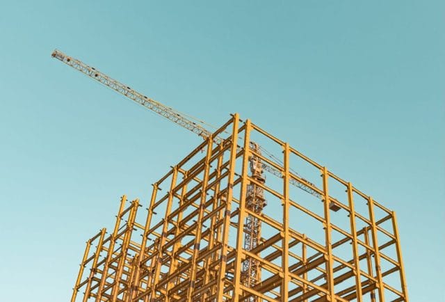A partially constructed building framework with yellow steel beams and a crane against a clear blue sky.