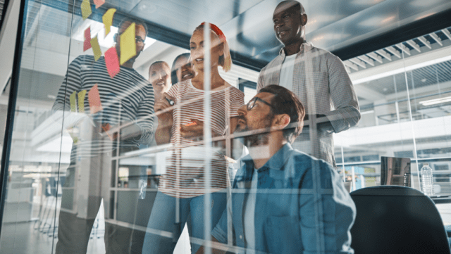 A diverse group of five people collaborates around a glass wall covered in colorful sticky notes, discussing ideas with one person holding a smartphone.
