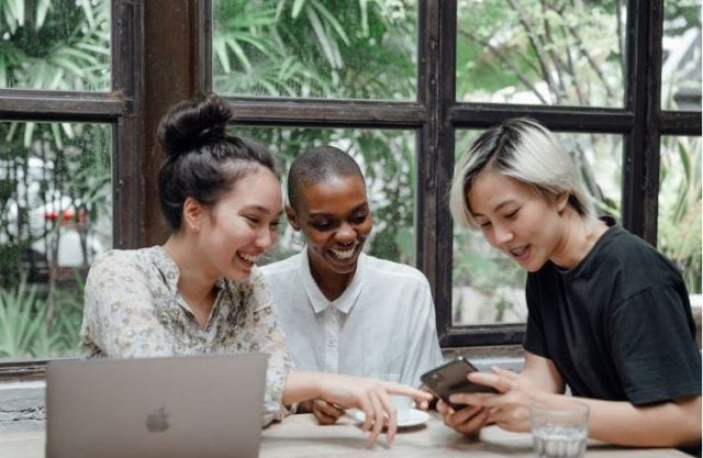 Three women sit at a table engaged in conversation, sharing a smartphone. A laptop is open in front of them, with green plants visible through large windows behind them.