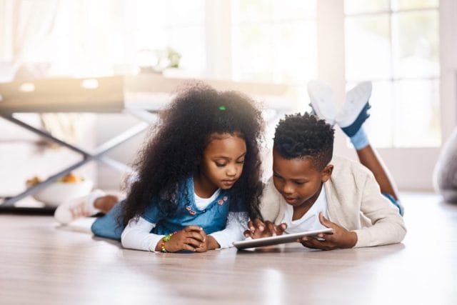 Two children sit on a wooden floor, focused on a tablet. One has curly hair and wears a blue shirt, while the other wears a light sweater. Natural light streams through a window behind them.