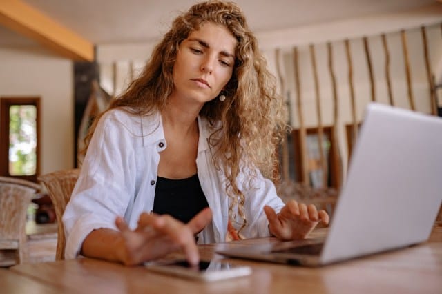 A focused woman with curly hair interacts with a laptop and smartphone on a wooden table, suggesting she is engaged in two-factor authentication for her business app.