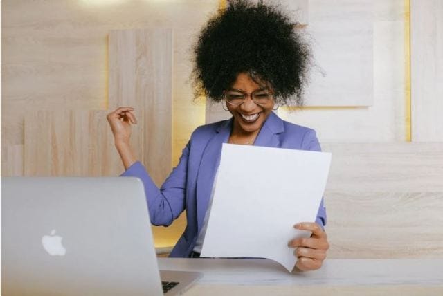 A woman in a blue blazer smiles while holding a sheet of paper and raising her hand joyfully. A laptop is visible on the table in front of her. The background features light wooden panels.