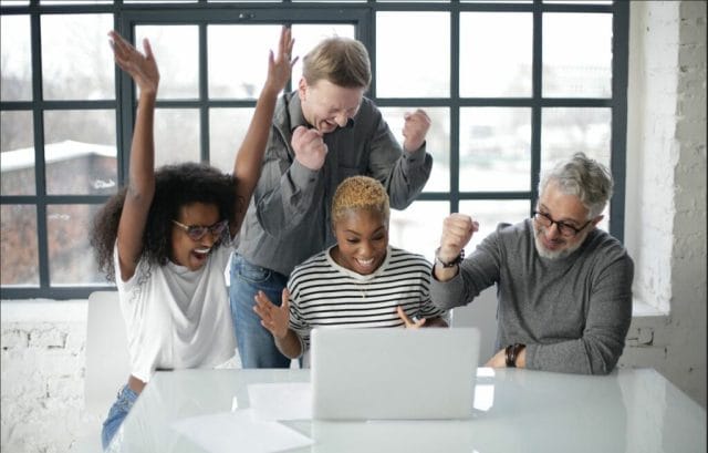 A diverse group of four people celebrating around a laptop at a table, showing excitement with raised arms and joyful expressions in a bright, modern workspace.