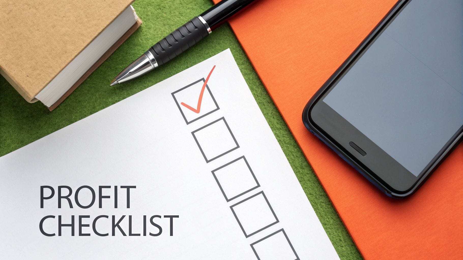 A checklist titled "Profit Checklist" with a checked box is displayed alongside a black pen, a closed tan notebook, and a smartphone, on a green felt surface with an orange folder.