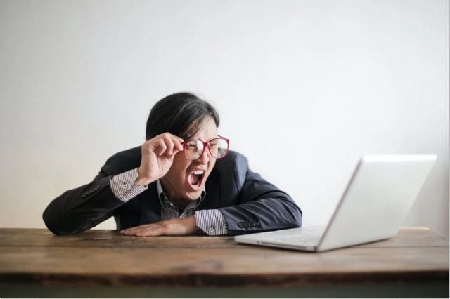 A person in a suit is leaning over a desk, looking frustrated while yelling at a laptop. They are wearing large red glasses and have a shocked expression. The background is plain white.